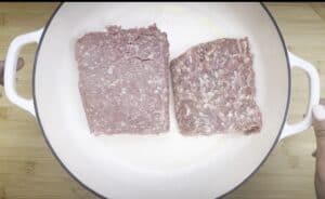 Two blocks of ground meat in a white pan on a wooden surface, ready for cooking.